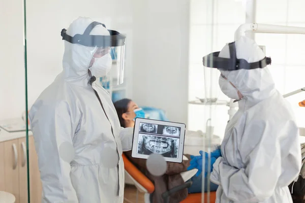 Médicos dentistas com traje ppe analisando raios-x dos dentes usando tablet — Fotografia de Stock