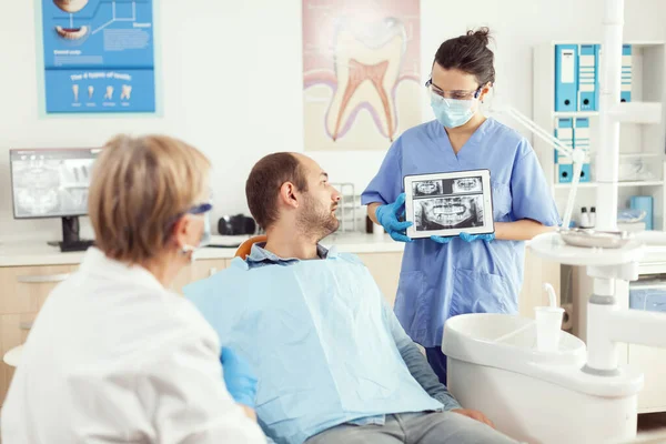 Женщина-дантист в стоматологическом кабинете разговаривает с больным мужчиной, пока медсестра показывает рентген зуба — стоковое фото