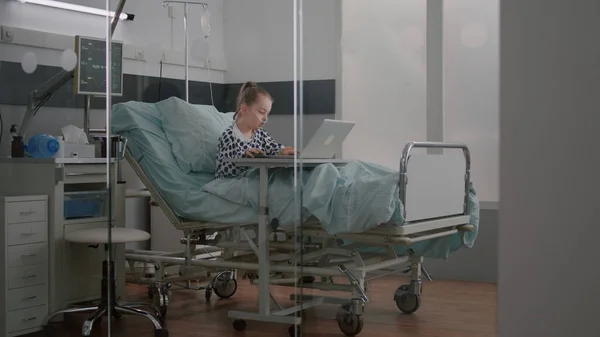 Criança doente relaxando na cama jogando videogames de desenhos animados no computador portátil durante o exame médico — Fotografia de Stock
