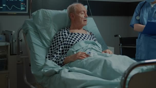 Медсестра проверяет дело больного пациента, лежащего в палате больницы — стоковое видео