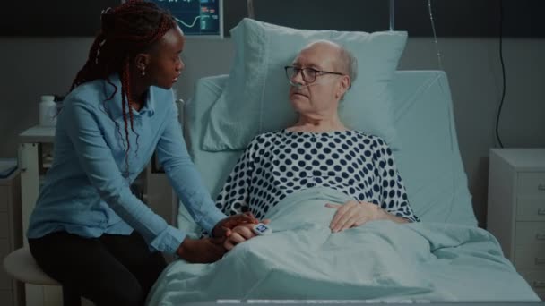 Больной пожилой пациент, лежащий в постели, разговаривает с африканским американским посетителем — стоковое видео