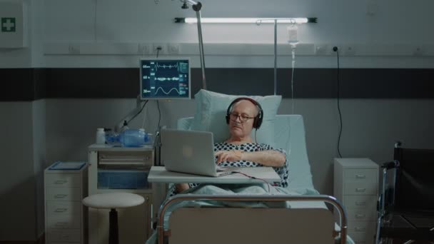 Un bătrân bolnav stând într-un spital folosind laptop și căști — Videoclip de stoc