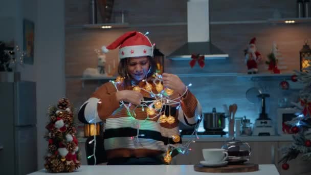 Glimlachende vrouw met kerstmuts verstrengeld in kerstverlichting — Stockvideo