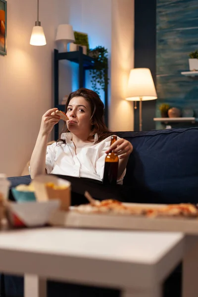 Кавказская женщина отдыхает на диване и ест пиццу, пьет пиво во время развлекательного фильма — стоковое фото