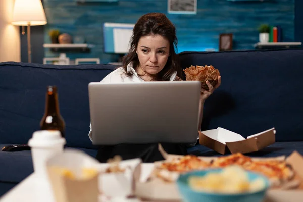 Mujer caucásica sentada en el sofá comiendo deliciosa hamburguesa sabrosa mientras trabaja en el ordenador portátil — Foto de Stock