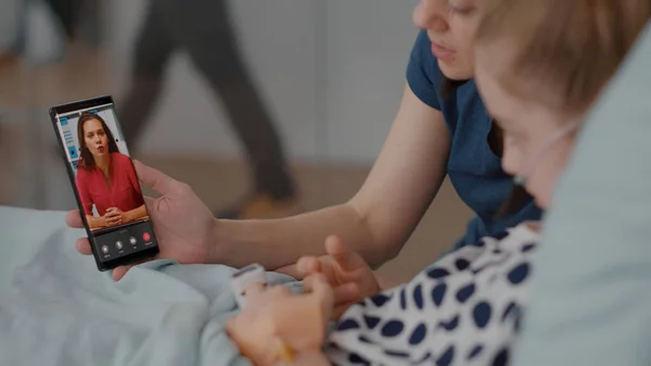 Больной пациент отдыхает в постели с матерью приветствуя удаленного друга во время онлайн видеоконференции — стоковое фото