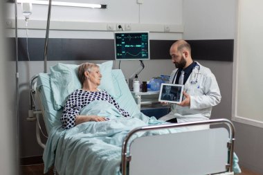 Yaşlı adamın yanında oturan doktor, vücut travması teşhisini açıklıyor.