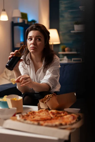 Mujer sosteniendo botella de cerveza relajándose en el sofá en la sala de estar viendo series documentales —  Fotos de Stock