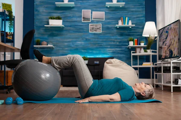 Aposentado sênior mulher fazendo praticar pernas até exercício usando suíço bola sentado no tapete de ioga — Fotografia de Stock