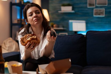 Beyaz kadın elinde lezzetli bir burgerle uzaktan komedi dizisi izleyerek kanal değiştiriyor.