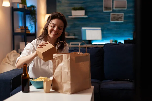 Lachende vrolijke vrouw uitpakken proeven fast-food huis geleverd zittend op de bank — Stockfoto