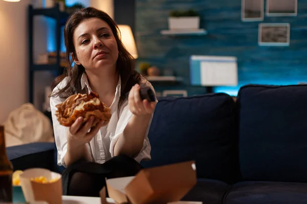 Blank vrouw holding smakelijke hamburger in handen veranderen kanalen met behulp van remote watching comedy serie — Stockfoto