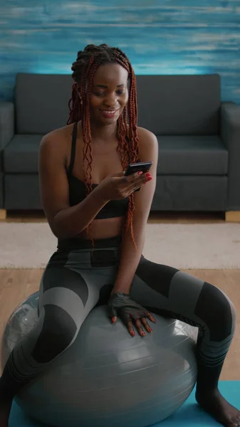 Slim fit mujer negra sentada en la pelota de fitness escribiendo mensaje en el teléfono —  Fotos de Stock
