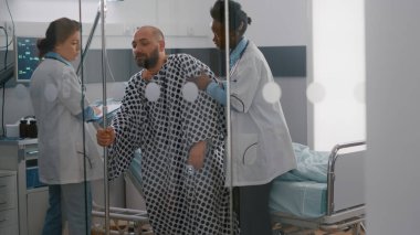 Pratisyen doktorlar hastaya yataktan kalkması için yardım ediyor. Damardan damar içi sıvı torbası tutuyorlar.