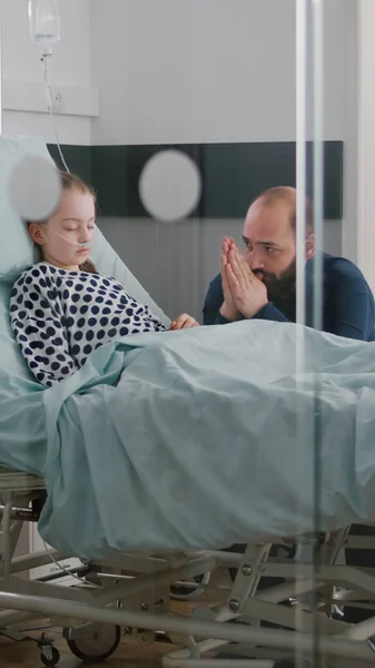 En la sala del hospital enferma niña paciente durmiendo mientras padre preocupado rezando por la recuperación de la enfermedad —  Fotos de Stock