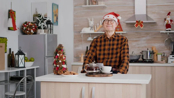 Retrato do avô vestindo chapéu de Papai Noel sentado em xmas cozinha decorada — Fotografia de Stock
