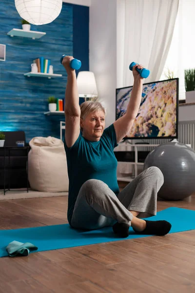 Retired senior woman sitting on yoga mat in lotus position raising hand during wellness routine — Stock Photo, Image