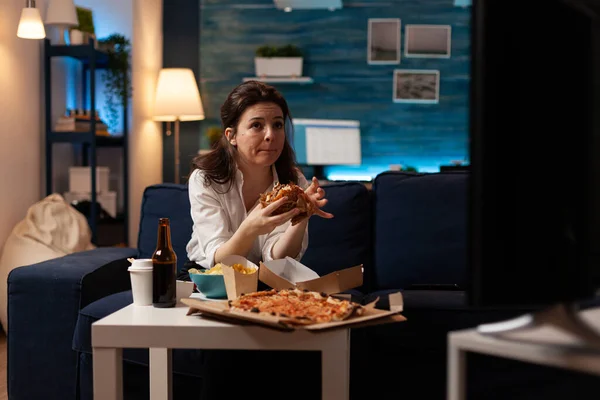 Woman sitting on sofa eating tasty delicious burger watching documentary movie