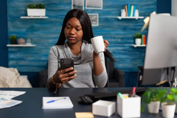 African american woman holding cup of coffee in hands socializing with friend using smartphone — ストック写真