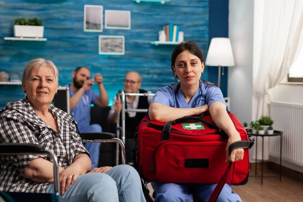 Портрет допоміжної медсестри, яка тримає набір ліків, сидить поруч з інвалідом старший пацієнт — стокове фото