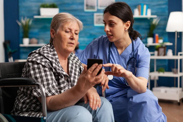 Enfermera ayudando a anciana jubilada en silla de ruedas a usar el teléfono inteligente durante el servicio social —  Fotos de Stock