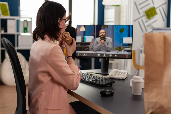 Empreendedor mulher sentada na mesa no escritório da empresa comendo sanduíche durante videocall conferência on-line — Fotografia de Stock