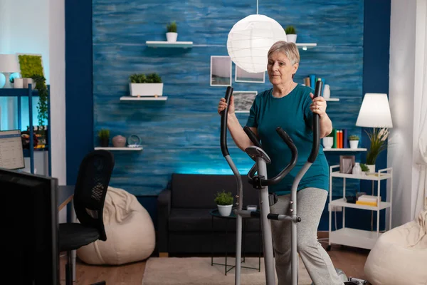 Senior woman doing aerobics on cycling bike machine in living room for well being — Stock Photo, Image