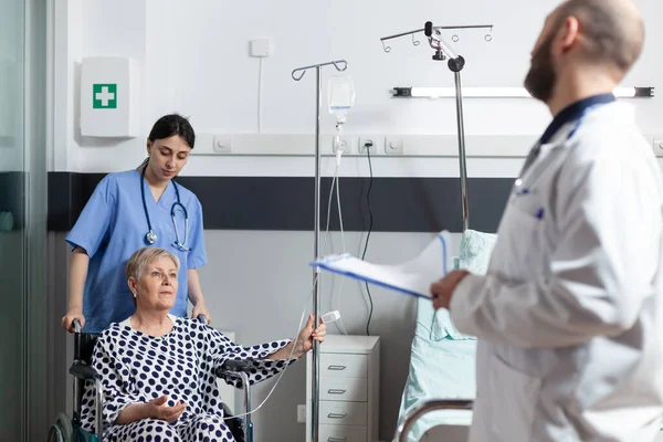 Zieke oudere vrouw zit in rolstoel met iv infuuszak bevestigd op arm — Stockfoto