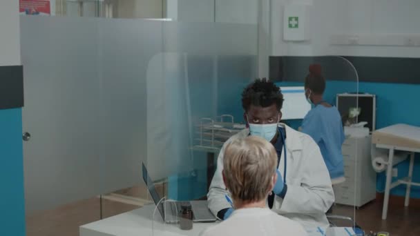 Medic with white coat sitting at desk while talking to elder woman — Stock Video