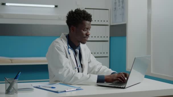 Afroamerykanski lekarz uzywajacy laptopa podczas siedzenia przy biurku — Wideo stockowe