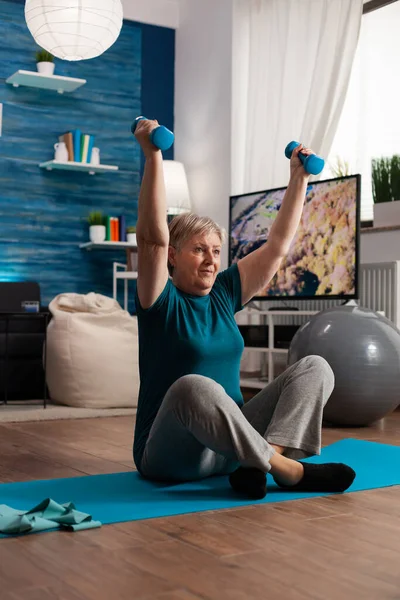 Retired senior woman sitting on yoga mat in lotus position raising hands streching arm muscle — Stock Photo, Image