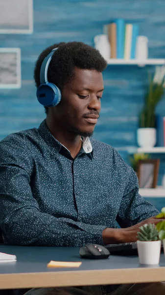 Gerente negro afroamericano usando auriculares para escuchar música mientras trabaja desde la oficina en el PC de la computadora —  Fotos de Stock