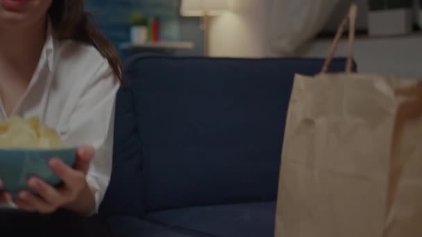 Portrait of young woman holding bowl of chips sitting on sofa — Stock Video