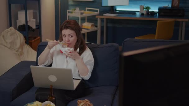 Young woman using laptop on sofa while eating asian food — Stock Video