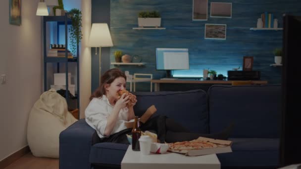 Молода жінка лежить на дивані їсть бургер і сміється — стокове відео
