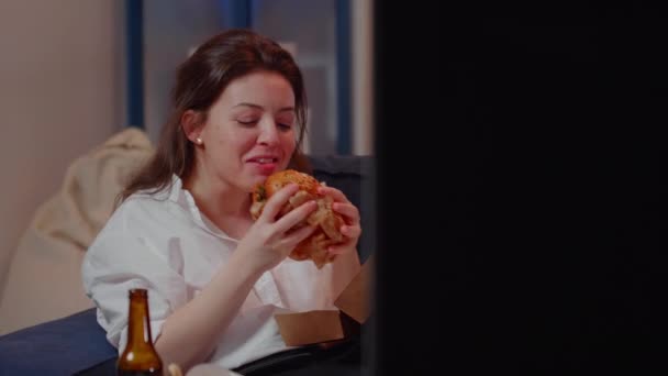 Zakelijke vrouw kijkt TV eten hamburger in de woonkamer — Stockvideo