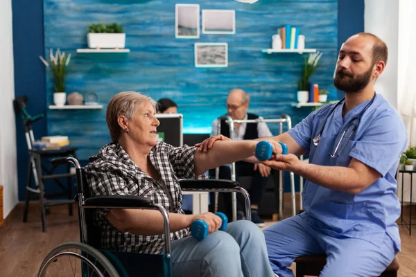 Чоловіча медсестра допомагає старшій жінці з інвалідністю у інвалідному візку реабілітуватися за допомогою гантелів — стокове фото