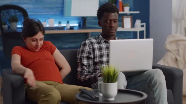 Mezirasový pár očekává dítě sedí spolu na gauči — Stock video