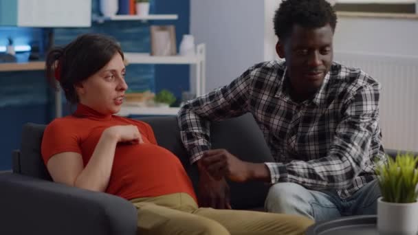 Hamil antar ras pasangan duduk di sofa ruang tamu — Stok Video