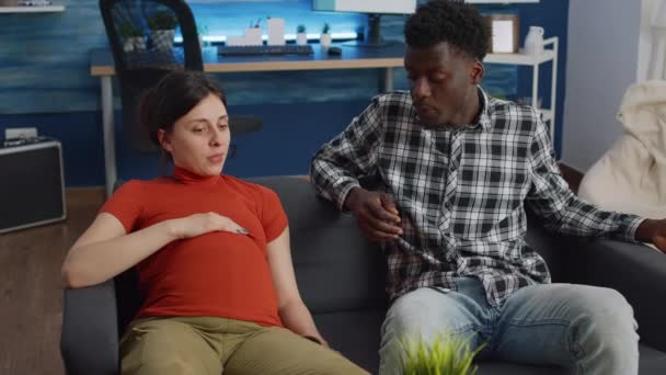 Embarazada interracial pareja sentado en sofá en casa — Vídeo de stock