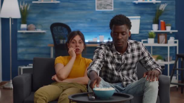 Молодая межрасовая пара смотрит телевизор и ест попкорн — стоковое видео