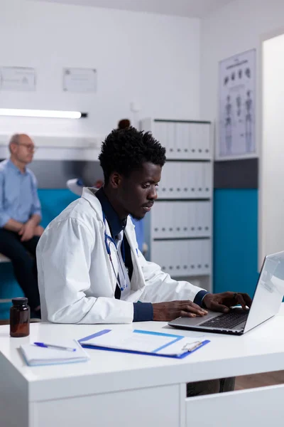 非洲裔美国医生坐在办公桌前使用笔记本电脑的画像 — 图库照片