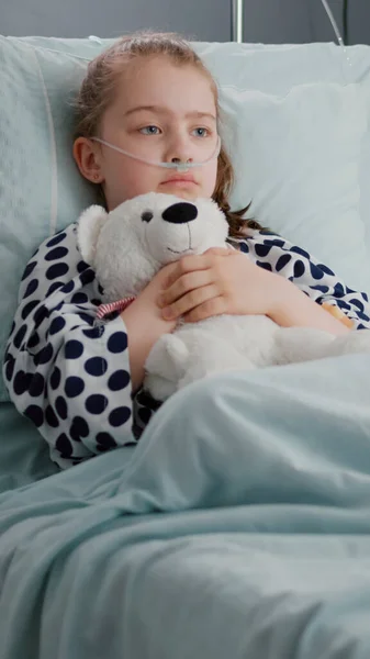 Один стурбований дитиною пацієнт носить кисневу носову трубку, яка спить у ліжку, тримаючи ведмедя-чаклуна — стокове фото