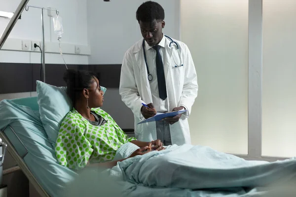 Disease doctor sitting with young afro patient in hospital ward — Stock Photo, Image