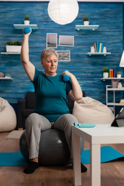 Senior woman in retirement raising hands doing arms exercise using dumbbells sitting fitball — Stock Photo, Image