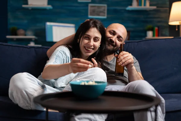 Casal animado em pijama refrigeração no sofá comer pipocas assistindo filme de comédia — Fotografia de Stock