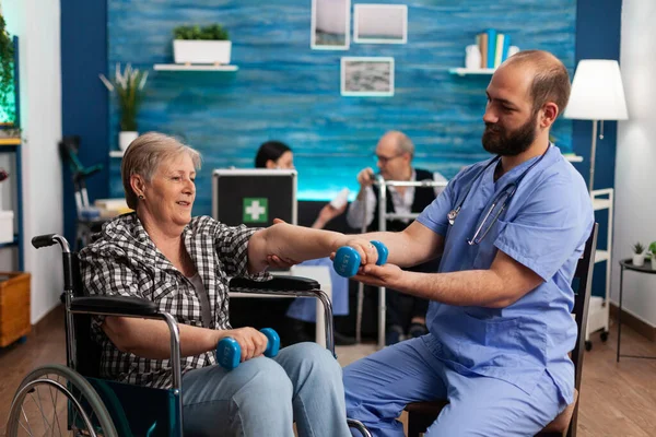 Чоловіча медсестра допомагає старшій жінці з інвалідністю у інвалідному візку реабілітуватися за допомогою гантелів — стокове фото