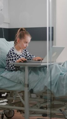 Hastanede oksijen tüpü takan hasta bir kız yatağında oturmuş dizüstü bilgisayarında video oyunları oynuyor.