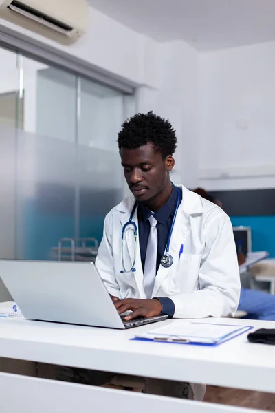 Retrato de médico afro-americano sentado na mesa usando laptop — Fotografia de Stock