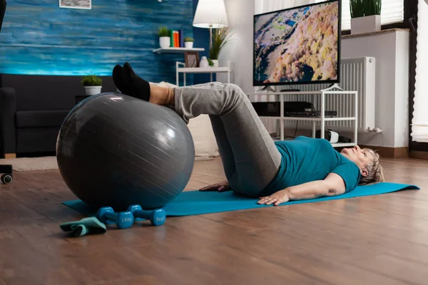 Gepensioneerde senior vrouw doet het beoefenen van benen omhoog oefening met behulp van Zwitserse bal zittend op yoga mat — Stockfoto